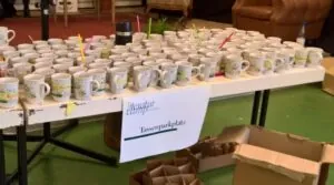 Ein Tisch auf dem ganz viele Tassen stehen, am Tischrand ist ein Plakat mit der Aufschrift "Tassenparkplatz" befestigt.