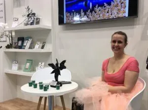 Ein Bild von Janna Ruth auf der Buchmesse: Sie sitzt rechts auf einem Stuhl und lacht in die Kamera, links ist ein kleiner, weißer Tisch, daneben ein Regal und an der Wand ein Bildschirm. 