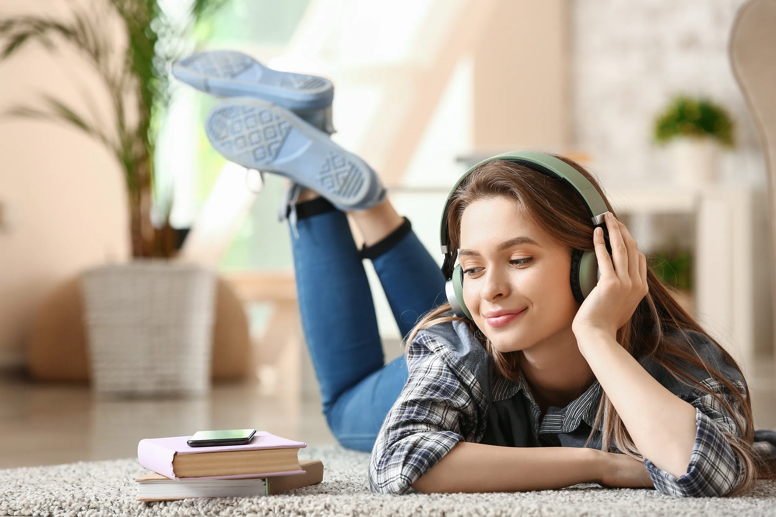 Beautiful young woman listening to audiobook at home Eine Frau liegt auf dem Boden, sie hat Kopfhörer auf und neben ihr liegen Bücher und ein Handy. Im Hintergrund sieht man eine Wohnung und eine Pflanze.