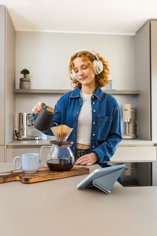 Foto Frau mit Kopfhörern Eine Frau steht mit Kopfhörern in einer Küche und bereitet sich einen Filterkaffe zu. Vor ihr steht ein Tablet auf dem Tisch.
